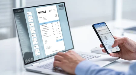 Ein Laptop mit einer geöffneten Rechnungsverwaltungssoftware zeigt eine digitale Rechnung. Eine Hand hält ein Smartphone mit einer Online-Banking-App.