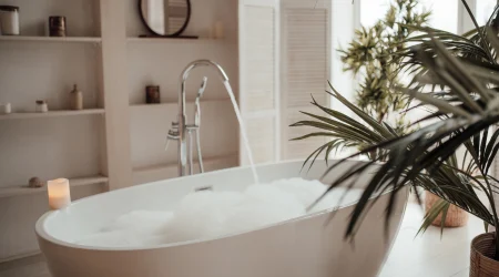 Badewanne gefüllt mit einem üppigen Schaumbad. Raumpflanzen ragen auf der rechten Seite in das Bild. Im Hintergrund ist ein minimalistisch gefülltes Regal zu sehen.
