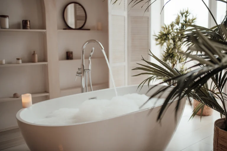 Badewanne gefüllt mit einem üppigen Schaumbad. Raumpflanzen ragen auf der rechten Seite in das Bild. Im Hintergrund ist ein minimalistisch gefülltes Regal zu sehen.