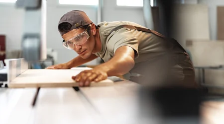 Schreiner mit Cap und Schutzbrille, schiebt ein Stück Holz durch eine Kreissäge.