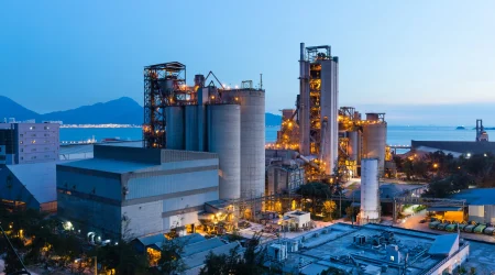 Industrielle Produktionsanlage mit Silos, Rohrsystemen und Türmen bei Dämmerung, beleuchtet und in Meeresnähe gelegen, im Hintergrund Berge und Wasser.