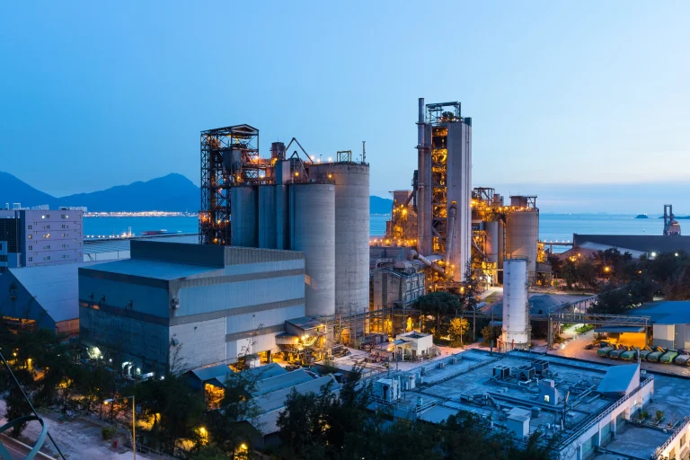 Industrielle Produktionsanlage mit Silos, Rohrsystemen und Türmen bei Dämmerung, beleuchtet und in Meeresnähe gelegen, im Hintergrund Berge und Wasser.