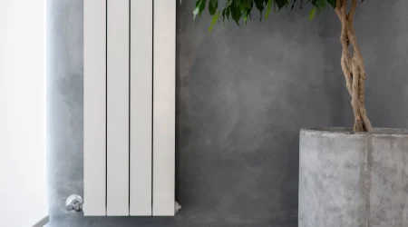 Weißer Niedertemperatur-Heizkörper mit vertikalen Paneelen, montiert an einer grauen Betonwand, daneben ein großer Beton-Pflanzkübel mit Baum.