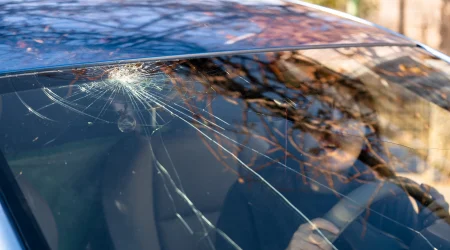 Steinschlag in einer Windschutzscheibe. Eine erschrockene Frau sitzt im Auto, hält sich am Lenkrad fest und schaut den Steinschlag an.