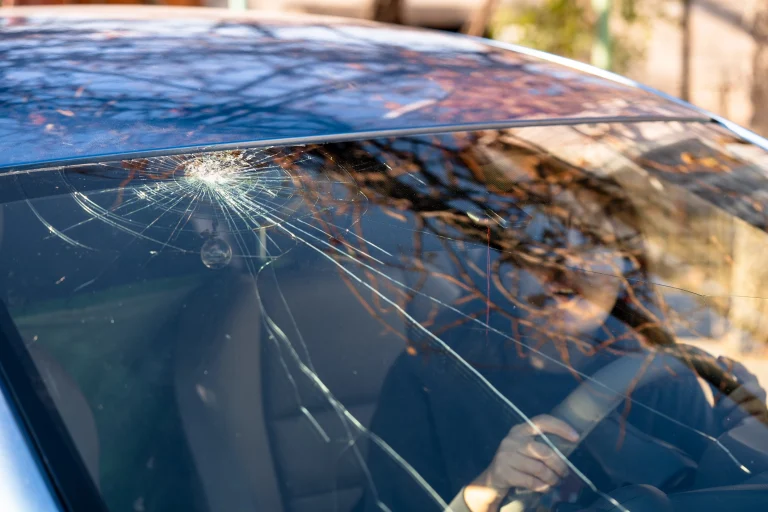 Steinschlag in einer Windschutzscheibe. Eine erschrockene Frau sitzt im Auto, hält sich am Lenkrad fest und schaut den Steinschlag an.