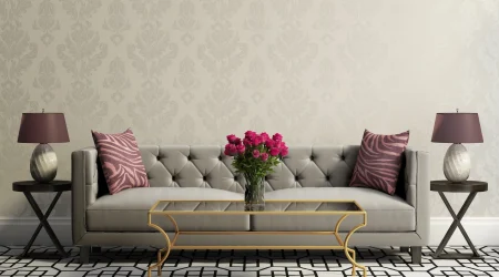 Ein stilvoll eingerichtetes Wohnzimmer mit einer eleganten Textiltapete in dezenten, floralen Muster. Im Mittelpunkt steht ein graues, gepolstertes Sofa mit zwei Zierkissen in lila Zebramuster. Vor dem Sofa steht ein goldener Couchtisch mit einer Glasplatte, darauf eine Vase mit frischen roten Rosen.
