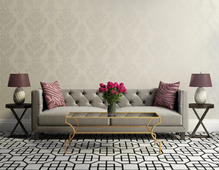 Ein stilvoll eingerichtetes Wohnzimmer mit einer eleganten Textiltapete in dezenten, floralen Muster. Im Mittelpunkt steht ein graues, gepolstertes Sofa mit zwei Zierkissen in lila Zebramuster. Vor dem Sofa steht ein goldener Couchtisch mit einer Glasplatte, darauf eine Vase mit frischen roten Rosen.