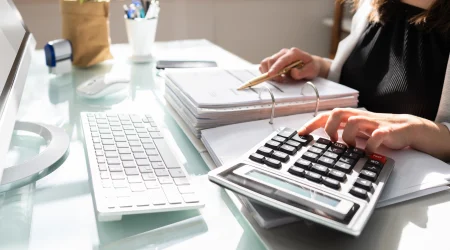 Eine Frau arbeitet an einem Schreibtisch mit Finanzunterlagen. Sie benutzt einen Taschenrechner und überprüft dabei Dokumente in einem Aktenordner mit einem Stift in der Hand. Auf dem Monitor ihres Computers ist eine Tabelle mit Rechnungsdetails zu sehen.
