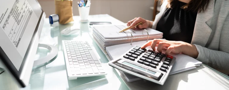 Eine Frau arbeitet an einem Schreibtisch mit Finanzunterlagen. Sie benutzt einen Taschenrechner und überprüft dabei Dokumente in einem Aktenordner mit einem Stift in der Hand. Auf dem Monitor ihres Computers ist eine Tabelle mit Rechnungsdetails zu sehen.