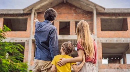 Eine kleine Familie bestehend aus Mutter, Vater und Kind stehen vor einem Rohbau eines Hauses. Der Vater zeigt seiner Familie etwas, indem er mit seinem Finger in die obere linke Ecke des Bildes deutet.