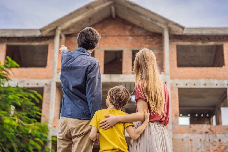 Eine kleine Familie bestehend aus Mutter, Vater und Kind stehen vor einem Rohbau eines Hauses. Der Vater zeigt seiner Familie etwas, indem er mit seinem Finger in die obere linke Ecke des Bildes deutet.