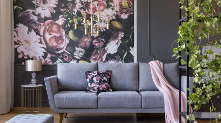 Modern eingerichtetes Wohnzimmer mit einer grauen Couch, auf der ein florales Kissen und eine rosa Decke liegen. Im Hintergrund eine große Fototapete mit einem Blumenmuster in Rosa-, Weiß- und Grüntönen. Links neben der Couch steht ein runder Beistelltisch mit einem goldenen Gestell und einer Tischlampe. Rechts im Vordergrund rankt eine grüne Pflanze an einem schwarzen Regal empor.