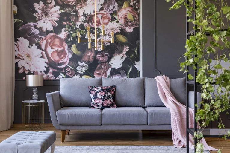Modern eingerichtetes Wohnzimmer mit einer grauen Couch, auf der ein florales Kissen und eine rosa Decke liegen. Im Hintergrund eine große Fototapete mit einem Blumenmuster in Rosa-, Weiß- und Grüntönen. Links neben der Couch steht ein runder Beistelltisch mit einem goldenen Gestell und einer Tischlampe. Rechts im Vordergrund rankt eine grüne Pflanze an einem schwarzen Regal empor.