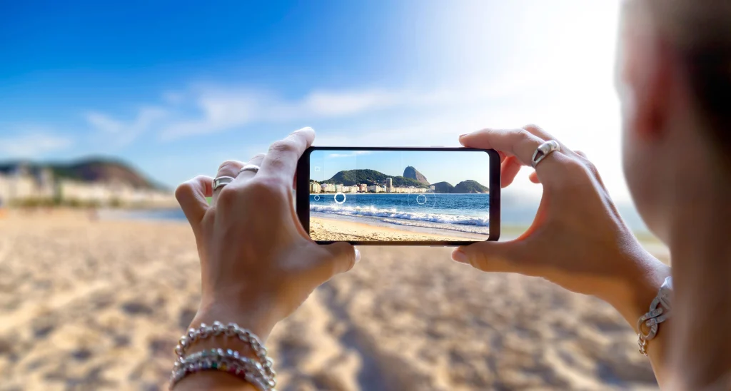 Eine Person hält ein Smartphone mit beiden Händen und fotografiert eine Strandlandschaft. Der Vordergrund mit den Händen, Ringen und Armbändern ist scharf, während der Hintergrund mit dem Strand, dem Meer und den Bergen unscharf ist. Auf dem Smartphone-Display ist die Landschaft hingegen klar und fokussiert zu sehen.