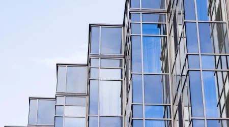Eine moderne Glasfassade aus Sonnenschutzglas mitversetzten, gestuften Fensterelementen. Die reflektierenden Glasflächen spiegeln den blauen Himmel und umgebende Gebäude wider, während die Metallrahmen eine geometrische Struktur bilden.