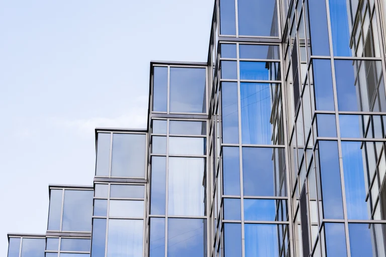 Eine moderne Glasfassade aus Sonnenschutzglas mitversetzten, gestuften Fensterelementen. Die reflektierenden Glasflächen spiegeln den blauen Himmel und umgebende Gebäude wider, während die Metallrahmen eine geometrische Struktur bilden.