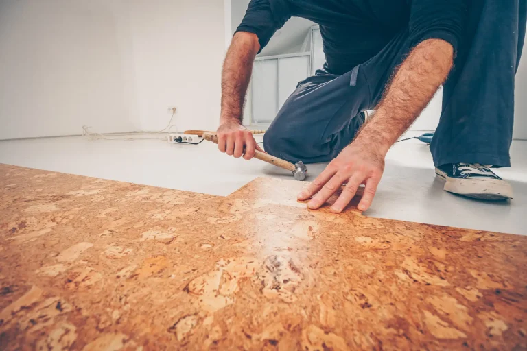 Ein Bodenleger in dunkler Kleidung kniet auf dem Boden und verlegt Korkboden. Er hält einen Hammer in der Hand und passt eine Korkplatte an. Der Raum hat weiße Wände und einen hellen Untergrund, im Hintergrund liegt eine Steckdosenleiste mit Kabeln.