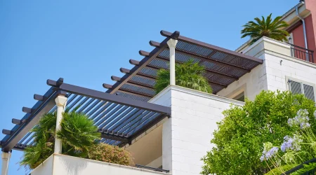 Moderne Pergola aus dunklem Metall mit Lamellen auf einem Balkon eines weißen Gebäudes. Die Pergola wird von hellen Säulen gestützt. Auf dem Balkon befinden sich grüne Palmen und weitere Pflanzen. Im Hintergrund ein blauer Himmel und ein Teil eines roten Gebäudes mit schwarzem Geländer.