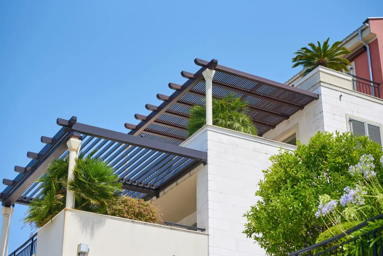 Moderne Pergola aus dunklem Metall mit Lamellen auf einem Balkon eines weißen Gebäudes. Die Pergola wird von hellen Säulen gestützt. Auf dem Balkon befinden sich grüne Palmen und weitere Pflanzen. Im Hintergrund ein blauer Himmel und ein Teil eines roten Gebäudes mit schwarzem Geländer.