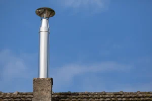 Schornsteinverlängerung aus Edelstahl mit Regenhaube auf einem gemauerten Schornsteinsockel, montiert auf einem mit Moos bedeckten Ziegeldach vor klarem, blauem Himmel.