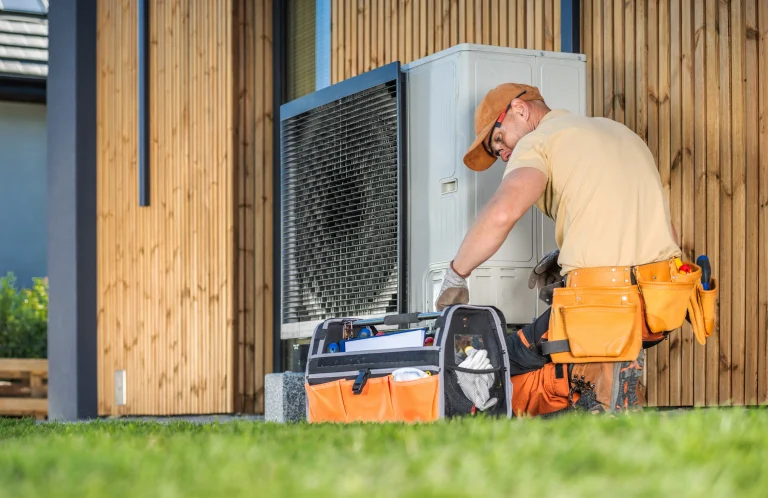 Ein Heizungstechniker in orangefarbener Arbeitskleidung kniet vor einer außen montierten Wärmepumpe und überprüft die Anlage. Daneben steht sein offener Werkzeugkoffer auf dem Rasen, während im Hintergrund eine Holzfassade zu sehen ist.