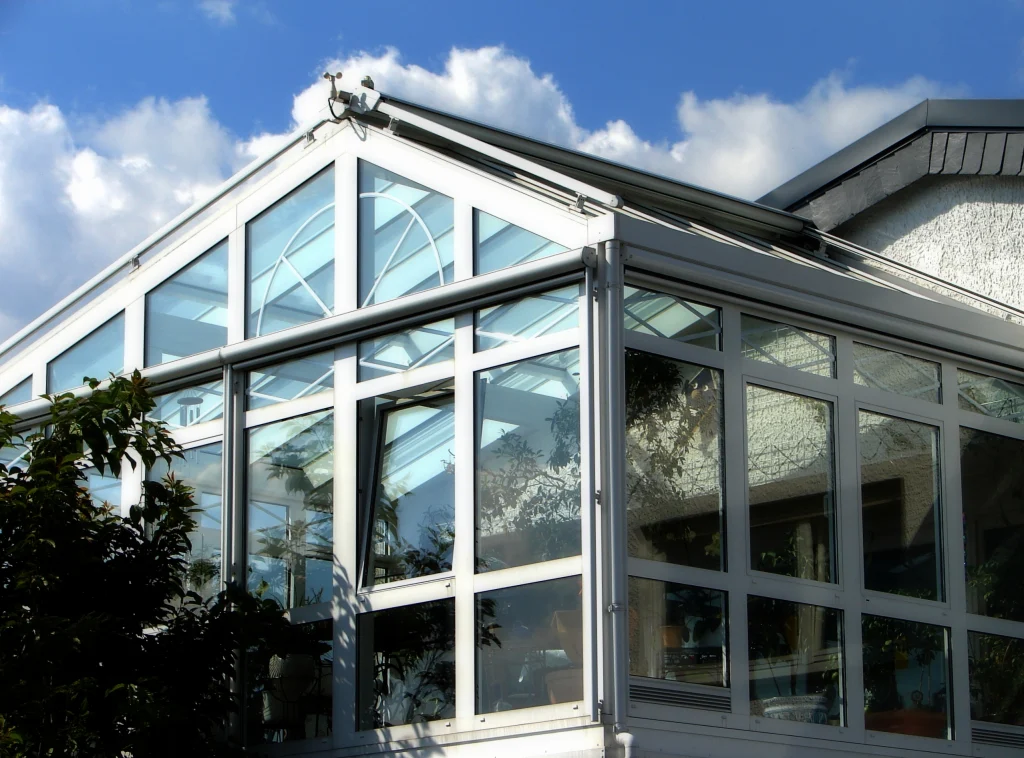 Ein moderner Wintergarten aus Aluminium mit großen Glasfenstern und einem spitz zulaufenden Dach. Die Konstruktion ist weiß und reflektiert den blauen Himmel mit einigen weißen Wolken. Im Inneren sind Pflanzen und Möbel zu erkennen, die eine gemütliche Atmosphäre schaffen. Ein Baum wirft seinen Schatten auf die Glasfront.