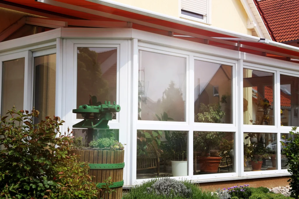 Ein Wintergarten mit weißem Kunststoffrahmen und großen Glasfenstern, der an ein Einfamilienhaus mit rotem Ziegeldach angrenzt. Im Inneren sind zahlreiche Topfpflanzen und Gartenmöbel sichtbar. Vor dem Wintergarten befindet sich ein bepflanztes Holzfass mit einer grünen Metallskulptur darauf, umgeben von Blumen und Sträuchern. Ein orangefarbenes Sonnendach ragt über den Wintergarten. Im Hintergrund sind Nachbarhäuser mit ähnlichen Dächern zu sehen.