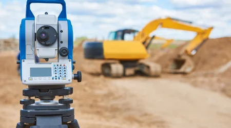 Vermessungsgerät steht zur Nutzung bereit auf einer Baustelle, im Hintergrund ist ein gelber Bagger zu sehen.
