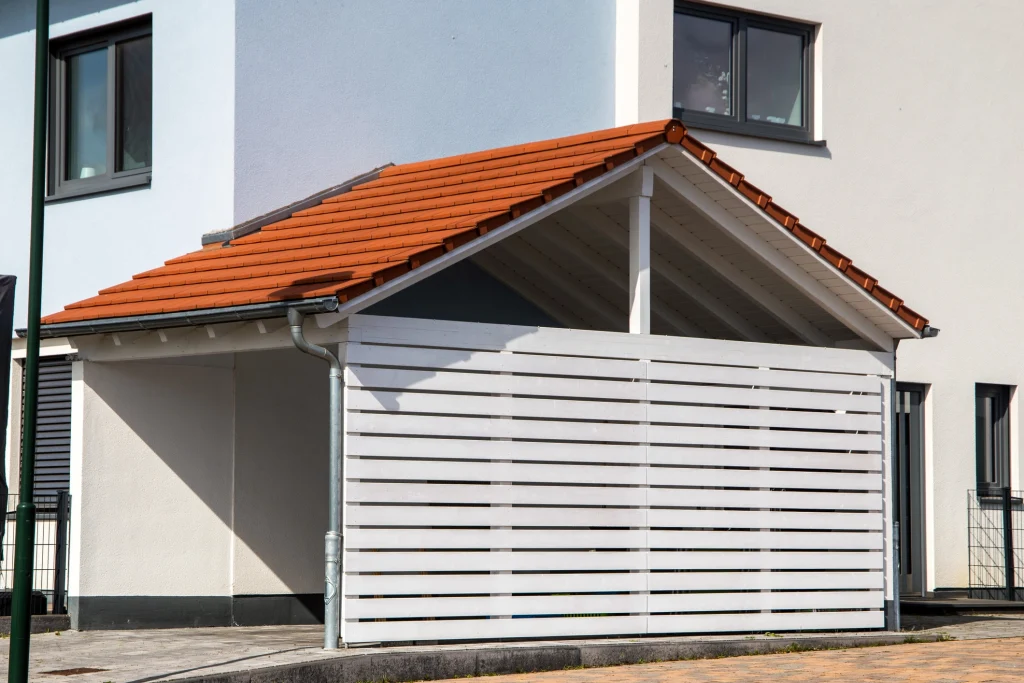 Ein moderner Carport mit einem roten Satteldach aus Ziegeln und einer weißen Holzverkleidung mit horizontalen Lamellen. Der Carport ist an ein weiß verputztes Wohnhaus angebaut und verfügt über eine Regenrinne mit Fallrohr. Im Hintergrund sind Fenster des Hauses sowie ein gepflasterter Bereich zu sehen.