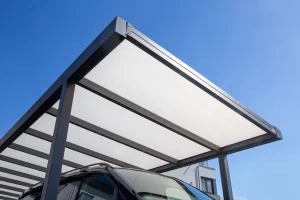 Modernes Carport mit lichtdurchlässigem Polycarbonatdach auf schwarzem Metallrahmen, unter dem ein schwarzes Auto im Schatten steht.