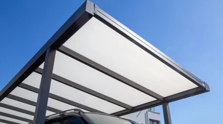 Modernes Carport mit lichtdurchlässigem Polycarbonatdach auf schwarzem Metallrahmen, unter dem ein schwarzes Auto im Schatten steht.