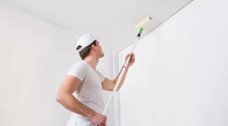 Maler in weißer Arbeitskleidung und Kappe tapeziert die Decke eines hellen Raumes. Er nutzt eine Teleskopstange, um die Tapete präzise anzubringen. Die Wand und Decke werden neu renoviert.