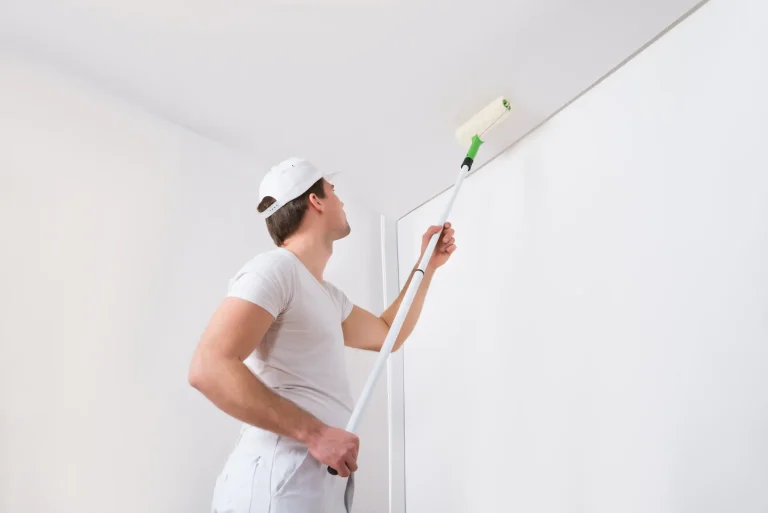 Maler in weißer Arbeitskleidung und Kappe tapeziert die Decke eines hellen Raumes. Er nutzt eine Teleskopstange, um die Tapete präzise anzubringen. Die Wand und Decke werden neu renoviert.