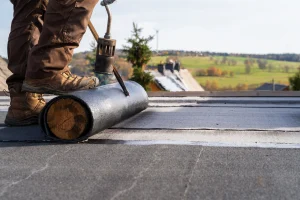 Ein Handwerker mit braunen Arbeitsschuhen und Hose verlegt zur Sanierung eine Bitumenbahn auf einem Flachdach. Mit einem Gasbrenner wird die Abdichtung erhitzt und auf dem Dach befestigt. Im Hintergrund ist eine ländliche Landschaft mit grünen Wiesen und Häusern mit Schornsteinen zu sehen.
