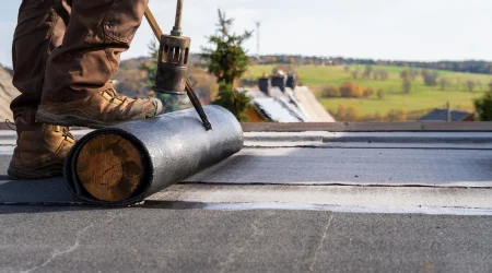 Ein Handwerker mit braunen Arbeitsschuhen und Hose verlegt zur Sanierung eine Bitumenbahn auf einem Flachdach. Mit einem Gasbrenner wird die Abdichtung erhitzt und auf dem Dach befestigt. Im Hintergrund ist eine ländliche Landschaft mit grünen Wiesen und Häusern mit Schornsteinen zu sehen.