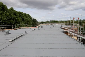 Sanierung eines Flachdachs mit neuer Abdichtung und umlaufendem Gerüst. Das Dach ist mit einer Bitumen- oder Kunststoffbahn gedeckt, während verschiedene Lüftungsrohre und technische Installationen sichtbar sind. Im Hintergrund sind Bäume und ein bewölkter Himmel zu sehen.