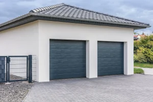 Moderne Doppelgarage mit einem Satteldach, gedeckt mit dunklen Dachziegeln. Die Fassade ist weiß verputzt, und die beiden Garagentore sind dunkelgrau. Vor der Garage befindet sich eine gepflasterte Einfahrt, und an der Seite steht ein Metallzaun mit Tor. Im Hintergrund sind einige Büsche und Häuser zu sehen.