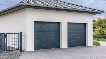 Moderne Doppelgarage mit einem Satteldach, gedeckt mit dunklen Dachziegeln. Die Fassade ist weiß verputzt, und die beiden Garagentore sind dunkelgrau. Vor der Garage befindet sich eine gepflasterte Einfahrt, und an der Seite steht ein Metallzaun mit Tor. Im Hintergrund sind einige Büsche und Häuser zu sehen.