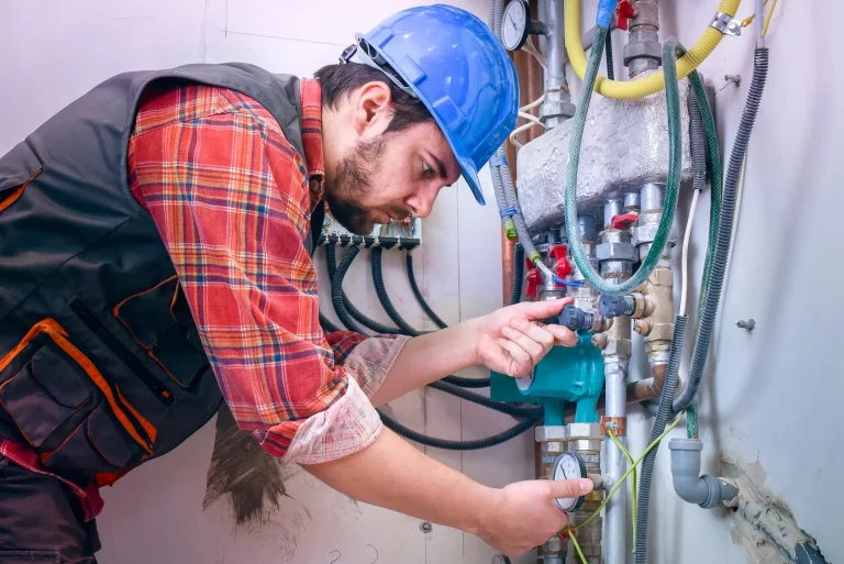 Handwerker mit blauem Schutzhelm und kariertem Hemd arbeitet an einer Heizungsanlage. Er überprüft Rohre, Ventile und ein Manometer. Im Hintergrund sind elektrische Verkabelungen und Isolierungen zu sehen.