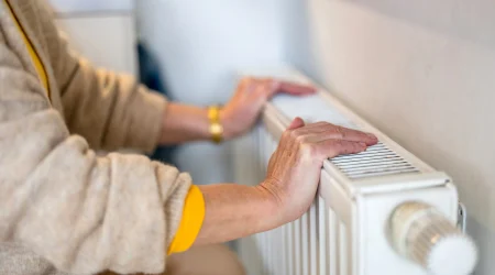 Eine Person prüft mit den Händen einen Heizkörper, der nur im oberen Bereich warm wird. Die Hände liegen auf der Oberseite des Heizkörpers, während der untere Teil offenbar kalt bleibt.