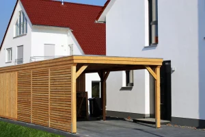 Ein moderner Carport aus Holz mit Flachdach steht neben einem weißen Wohnhaus mit roten Dachziegeln. Die Seitenwände des Carports bestehen aus horizontalen Holzlatten, die teilweise geschlossen sind. Der Boden ist mit dunklen Pflastersteinen ausgelegt, und im Hintergrund sind Fenster und Türen des Hauses sichtbar.