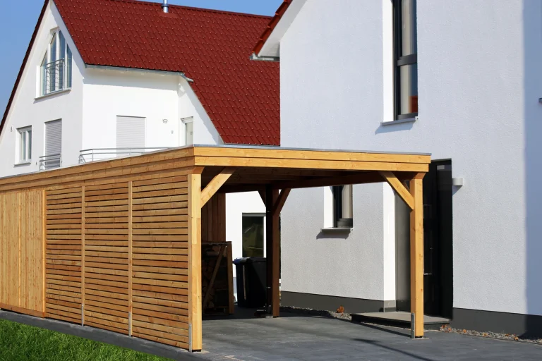Ein moderner Carport aus Holz mit Flachdach steht neben einem weißen Wohnhaus mit roten Dachziegeln. Die Seitenwände des Carports bestehen aus horizontalen Holzlatten, die teilweise geschlossen sind. Der Boden ist mit dunklen Pflastersteinen ausgelegt, und im Hintergrund sind Fenster und Türen des Hauses sichtbar.