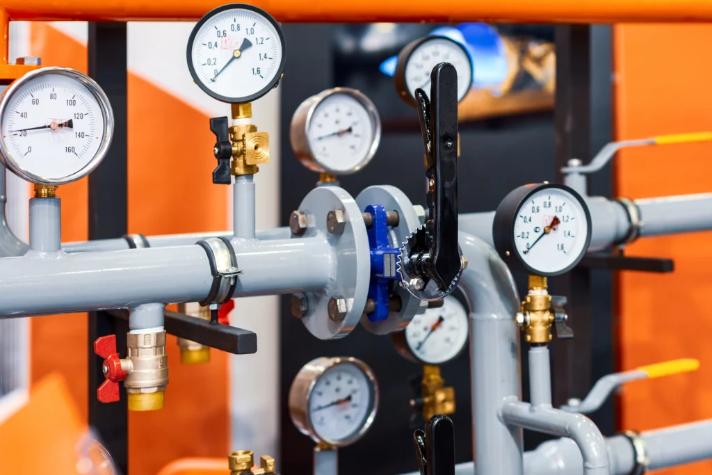 Detailaufnahme eines hydraulischen Systems mit mehreren grauen Rohrleitungen, analogen Druckmanometern und Ventilen. Die Druckanzeigen zeigen unterschiedliche Werte, und ein schwarzer Hebel sowie rote und gelbe Absperrventile sind sichtbar. Im Hintergrund befinden sich orangefarbene Wände.