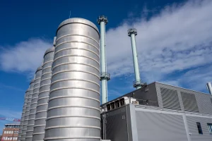 Moderne Kraft-Wärme-Kopplungsanlage mit mehreren silbernen, zylindrischen Speichertanks im Vordergrund und hohen, hellgrünen Abgasrohren im Hintergrund. Die Anlage besteht aus einem Gebäude mit metallischer Außenverkleidung unter blauem Himmel mit einigen Wolken.