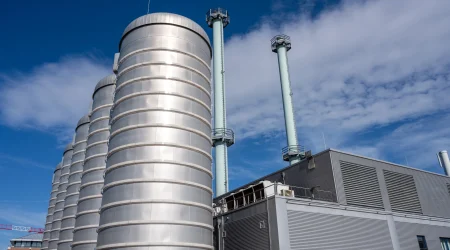 Moderne Kraft-Wärme-Kopplungsanlage mit mehreren silbernen, zylindrischen Speichertanks im Vordergrund und hohen, hellgrünen Abgasrohren im Hintergrund. Die Anlage besteht aus einem Gebäude mit metallischer Außenverkleidung unter blauem Himmel mit einigen Wolken.