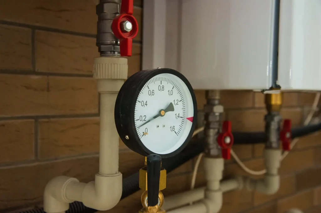 Nahaufnahme eines Manometers an einem Heizsystem mit weißen Rohren und einem roten Absperrventil. Das Druckmessgerät zeigt etwa 1,3 bar an. Im Hintergrund sind eine braune Kachelwand und weitere Rohrleitungen sichtbar.