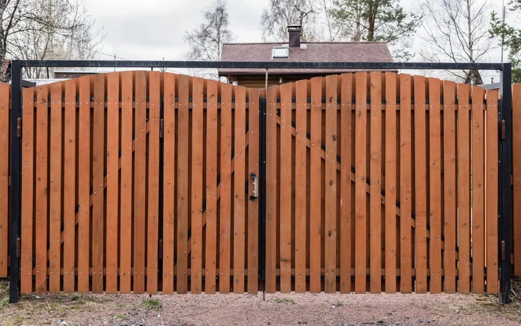 Ein großes zweiflügeliges Holztor mit schwarzem Metallrahmen, das den Eingang zu einem Grundstück mit einem Haus im Hintergrund sichert. Das Tor besteht aus senkrechten Holzlatten mit sichtbaren Schrauben und verstärkenden Metallstreben. Der Boden davor ist mit Kies bedeckt.