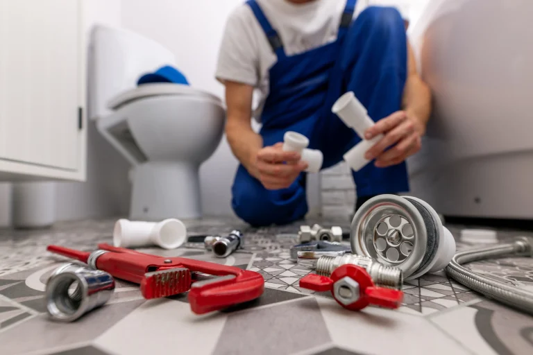 Eine Person in blauer Arbeitskleidung arbeitet in einem Badezimmer. Auf dem Boden liegen Werkzeuge und Sanitärteile, darunter eine rote Rohrzange, Rohre und Verschraubungen. Im Hintergrund ist eine Toilette zu sehen.