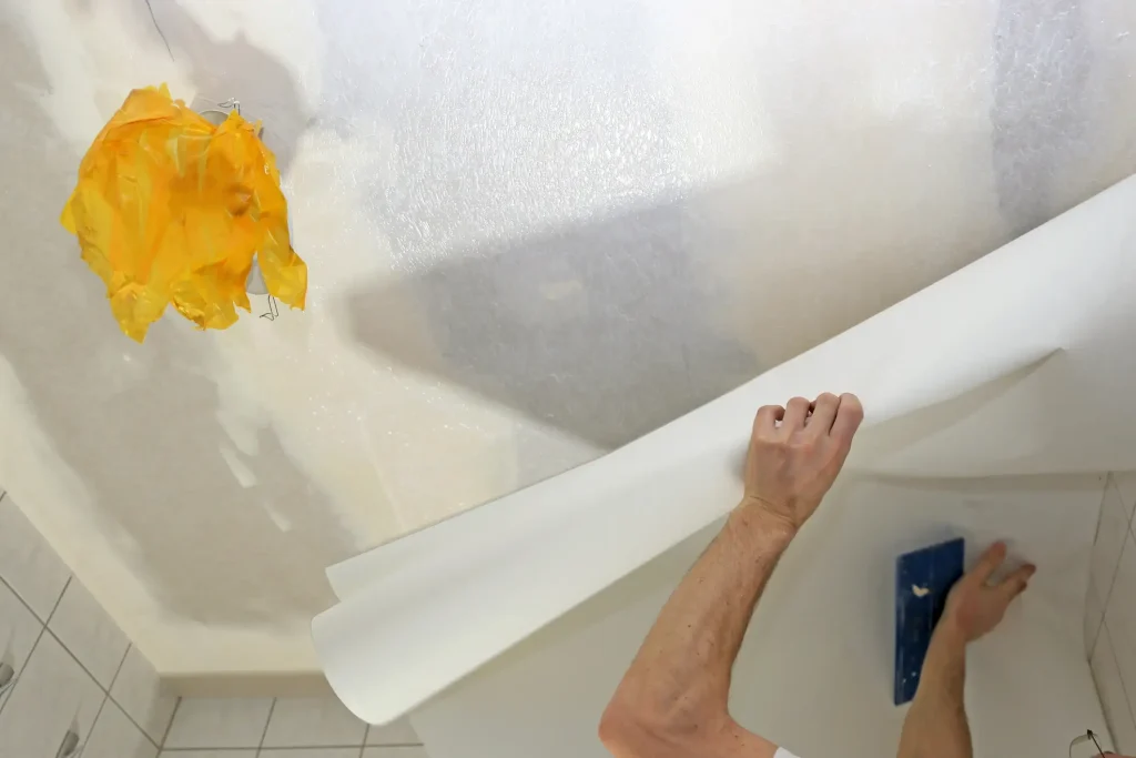 Handwerker tapeziert eine Decke mit weißer Tapete. Eine Hand hält die Tapetenbahn, während die andere mit einem Tapezierspachtel die Tapete an die feuchte Kleisterfläche andrückt. Eine mit gelbem Klebeband geschützte Lampenfassung ist an der Decke sichtbar.