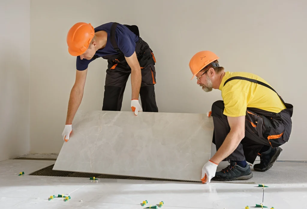 Zwei Handwerker mit orangefarbenen Schutzhelmen und Handschuhen verlegen eine große, helle Fliese auf einem Boden mit bereits ausgelegten Fliesennivellierern. Sie arbeiten sorgfältig und im Team, um die Fliese passgenau zu platzieren. Die Umgebung deutet auf eine Baustelle oder Renovierung hin, mit einer Fußbodenheizung, die unter den Fliesen verlegt sein könnte.